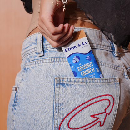 A close-up lifestyle shot of a person's hand holding a Chak and Go Coconut Crunch chocolate bar tucked into the back pocket of light-wash denim jeans with a red embroidered arrow.
